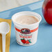 A white container of September Farm strawberry yogurt on a blue tray with a spoon and a red apple.