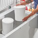 A person in a professional kitchen placing a white 3 gallon HDPE ice cream tub into a refrigerator.