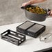 A person using an American Metalcraft black stainless steel crate to serve food from a pot on a white counter.