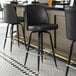 Three Lancaster Table & Seating black vinyl bucket seat cushions on black bar stools with metal legs at a bar counter.