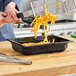 A person using tongs to serve french fries from a black Vollrath food pan.