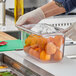 A person in gloves holding a Vollrath clear plastic food pan filled with oranges.