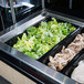 A Vollrath black polycarbonate food pan filled with chicken, lettuce, and other vegetables on a buffet counter.