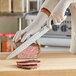 A person in gloves using a Choice Classic Granton Edge Pointed Carving Knife to cut meat on a wooden cutting board.