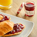 A croissant with Bonne Maman raspberry preserves on a plate next to more croissants and a jar of jam.