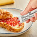 A hand holding a Bonne Maman Strawberry Preserves stick packet over a croissant.