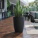 A Lancaster Table & Seating black planter with a potted plant on a wood floor.