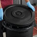 A person wearing blue gloves placing a black Advantage Supplies trash can lid on a black garbage can.