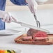 A person using an American Metalcraft stainless steel serrated knife to cut meat on a wooden plate.