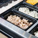 A Vollrath black polycarbonate food pan filled with food on a buffet counter.