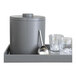 A grey room360 ice bucket with a metal lid on a tray with two glasses and a metal ladle.