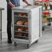 A man standing in front of a Regency stainless steel sheet pan rack with clear acrylic doors filled with bread.