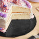 A cake with purple and white frosting on an Enjay black round cake drum with a slice cut out on a plate with a knife.