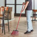 A man using an O-Cedar commercial broom with red bristles and a steel handle to sweep the floor.