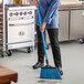 A woman in a chef's uniform using an O-Cedar blue angle broom with flagged bristles to sweep the floor in a professional kitchen.