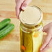 A hand holding a gold and white 82 metal lid over a jar of pickles.