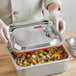 A person using a gloved hand to place a Vigor stainless steel cover on a metal container of food.