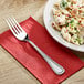 A plate of pasta on a table with a fork on a red napkin.