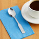 A Choice medium weight stainless steel teaspoon on a blue napkin next to a cup of coffee.