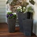Three Novelty Manufacturing Co. black Artstone planters on a table in a outdoor patio filled with purple flowers.