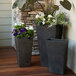 Three Novelty Manufacturing Co. black square planters on an outdoor table with purple flowers in them.