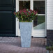 A Novelty Manufacturing Co. grey tall square planter with flowers in it.