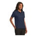 A woman wearing a navy Port Authority silk touch pique polo shirt.