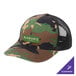 A Port Authority Woodland Camo and Black trucker cap with a white logo on the front.