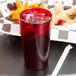 A Cambro ruby red plastic tumbler filled with a red drink and ice on a counter with a straw holder.