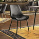 A Lancaster Table & Seating black vinyl chair with metal legs sits on a rug in front of a wooden table in a restaurant.