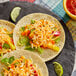 A group of three tacos with Plant Ahead vegan shredded cheese and vegetables on a black plate.