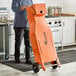 A person standing next to a large orange Fryclone 70-100 lb. Fryer Oil Shuttle on a counter in a professional kitchen.