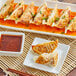 A plate of Chef One Pork and Napa Cabbage Dumplings with a bowl of red sauce and chopsticks.