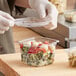 A person in gloves holding a Good Natured clear plastic deli container with food in it.