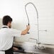 A man in a white shirt and black apron using a wall-mounted pre-rinse faucet over a sink.