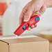 A hand using a red Allway Tools plastic vanishing knife to open a box.
