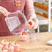 A woman in a pink apron holding a Good Natured clear plastic container with a cupcake with a strawberry on top.