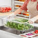 A woman in an apron holding a deep clear polycarbonate food pan with vegetables in it.