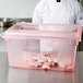 A person in a chef's uniform and white coat holding a red Carlisle plastic food storage container with meat in it.