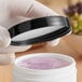 A hand in gloves holds a 70/400 smooth black lid over a white container of purple gel.