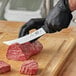 A person in black gloves using a Victorinox breaking knife to cut raw meat on a wooden cutting board.