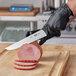 A person wearing black gloves using a Victorinox Churrasco Slicing Knife to cut meat on a cutting board.