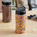 A round plastic jar full of colorful sprinkles with a black lid with 7 holes on a table.