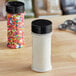 A glass container with white powder and colorful sprinkles in a round plastic spice container with a black lid with three holes.