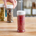 A hand holding a round white plastic spice container filled with red peppercorns.