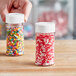 A hand holding a round plastic spice container filled with sprinkles on a table.