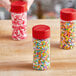 A group of 4 oz. round plastic spice containers with red flip and sift lids filled with colorful sprinkles.