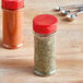 Two red round plastic spice containers with red dual flapper lids with 7 holes filled with spices.