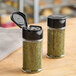 Two 3.5 oz. round plastic spice containers with black flip and sift lids filled with green herbs.