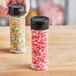 A hand holding a round plastic jar filled with colorful sprinkles with a black flip and sift lid.
